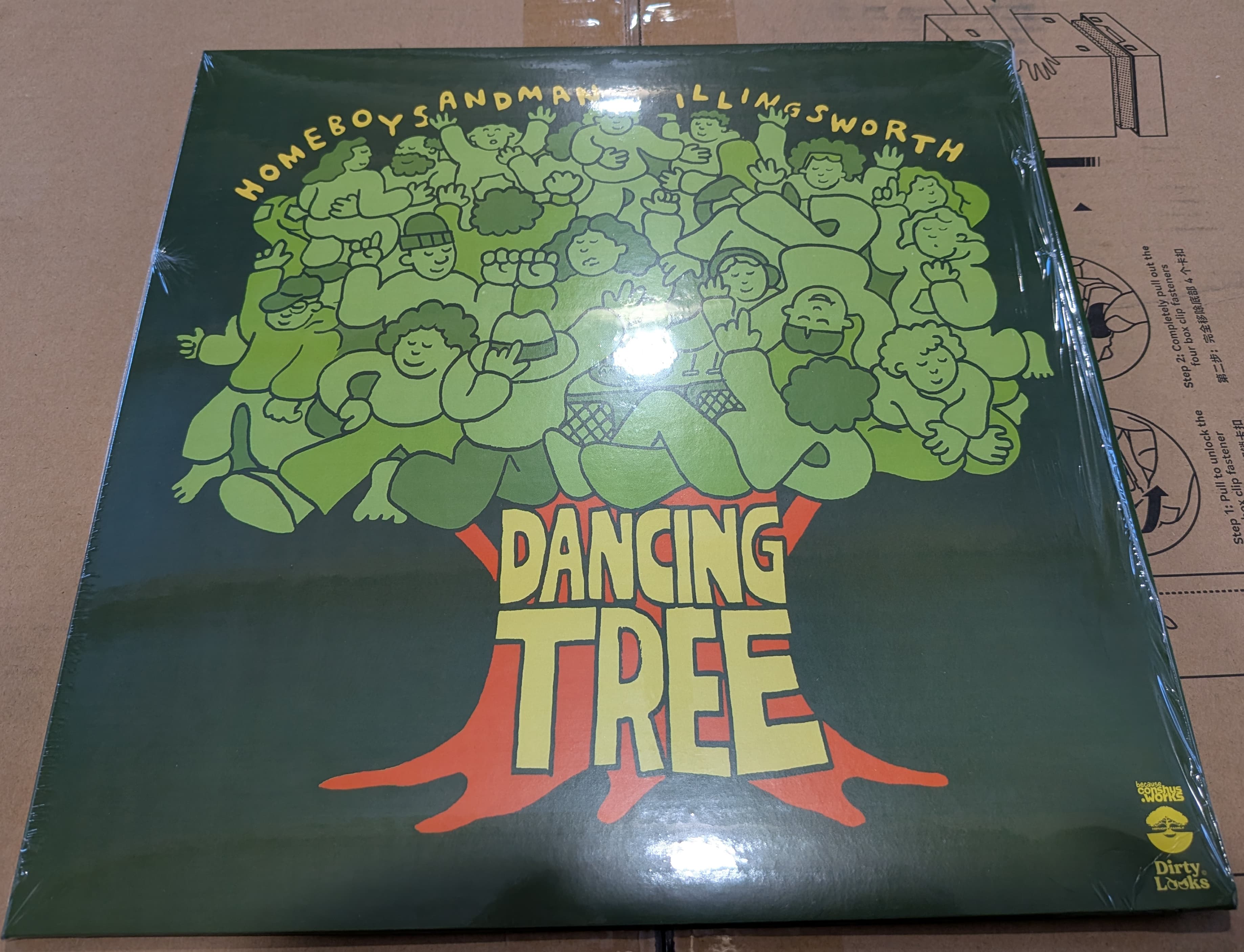 The album cover for 'HOMEBOYSANDMAN ILLINGSWORTH DANCING TREE' featuring a stylized illustration of numerous cartoonish figures with various expressions, drawn in shades of light and dark green, forming the leafy canopy of a tree. The tree's trunk and roots are depicted in orange with the words 'DANCING TREE' in large, yellow, stylized letters outlined in orange. The album title 'HOMEBOYSANDMAN ILLINGSWORTH' is written in a curved, yellow font at the top. The cover has a slightly glossy appearance, likely due to the plastic wrapping, and shows some reflections. In the bottom right corner, there are logos, one partially obscured by glare. The background is a slightly visible cardboard surface.